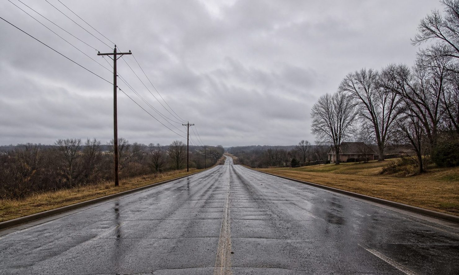 Wet two-lane road under a low grey sky, power lines and bare trees along the shoulder, outskirts of the St. Louis metro